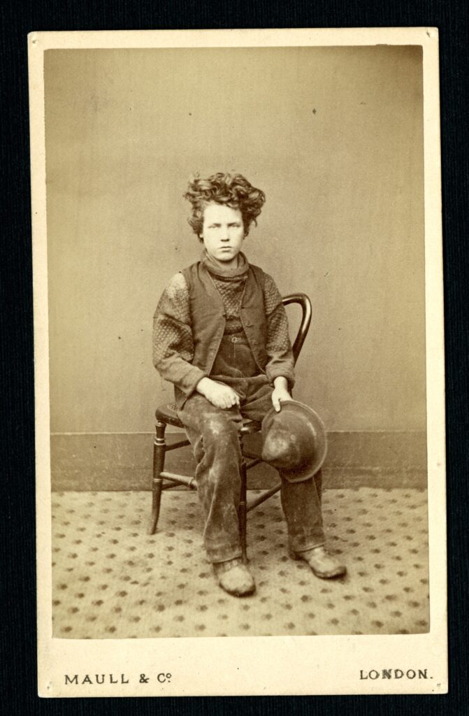 A white boy with tousled hair of about age ten sits in a chair, his hands on his knees looking slightly off camera. He wears a vest, neckerchief, and pants, and is holding a hat in his left hand.